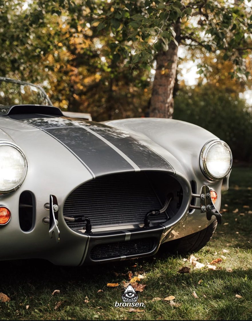 A silver Cobra roadster with racing stripes beneath autumn trees