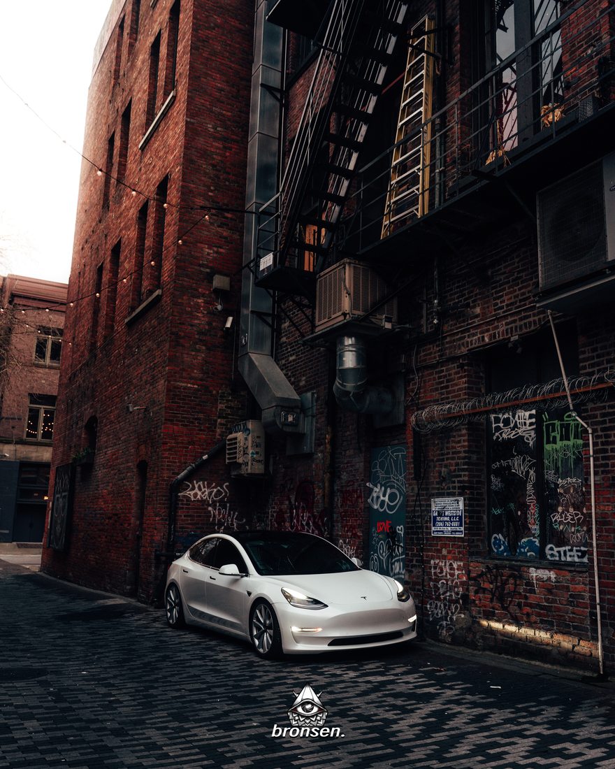 A white Tesla Model 3 parked in an old brick alley with fire escapes